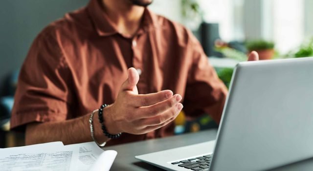 Student-taking-a-video-call-on-a-laptop-at-a-desk-covered-in-papers-scaled